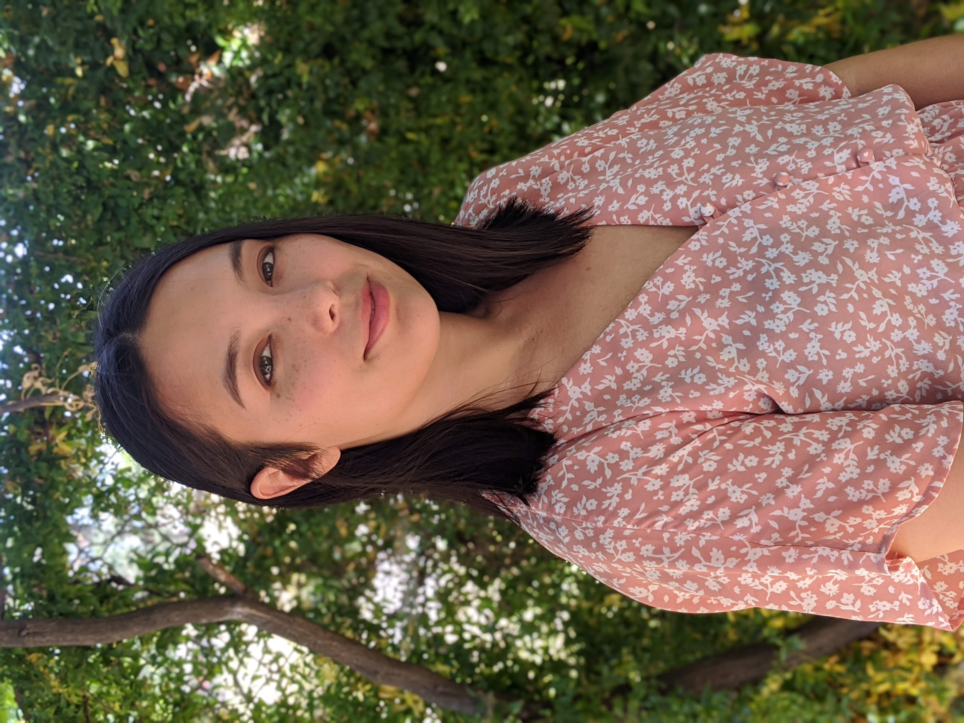 Young woman with dark shoulder length hair wearing a rose colored blouse, standing in front of green trees