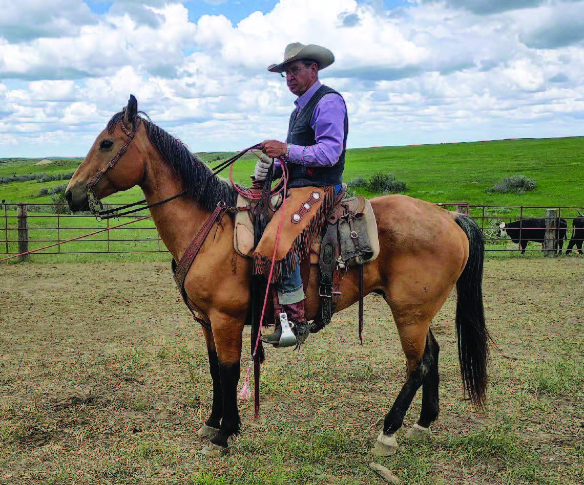 Jim sits atop a saddled horse, in chaps, holding a rope.