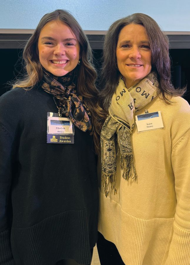 Two women, a mother and daughter, smile at the camera with their arms around each other's back. They are both wearing  name badges.