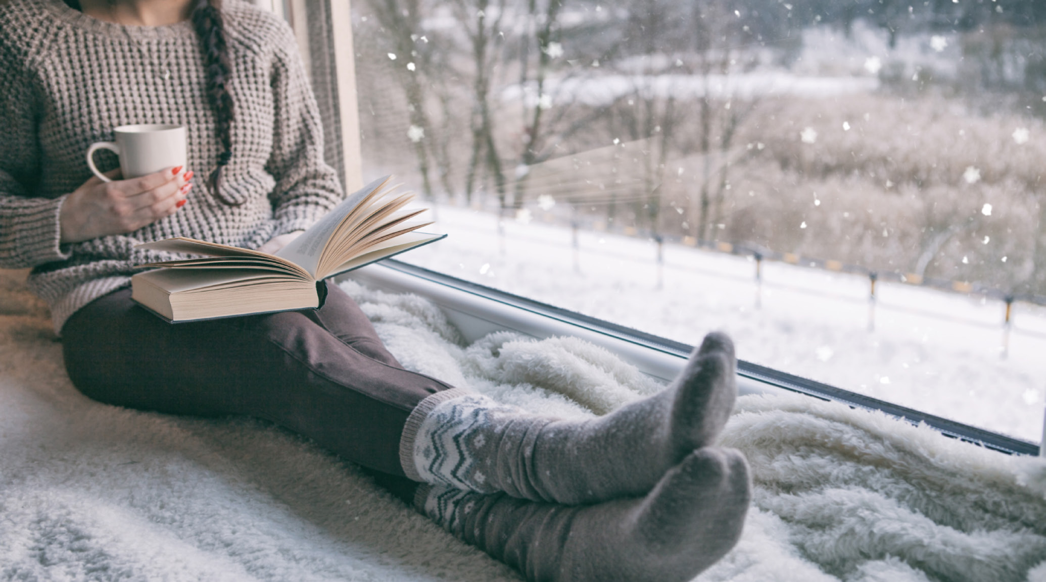 A woman sitting on a windowsill with a hot beverage and a book