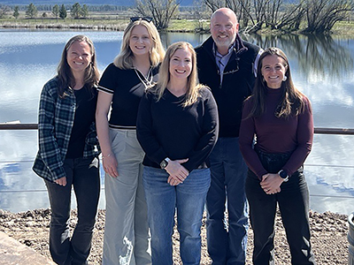 Four women and a man pose for a photo in front of a pond on a sunny day.