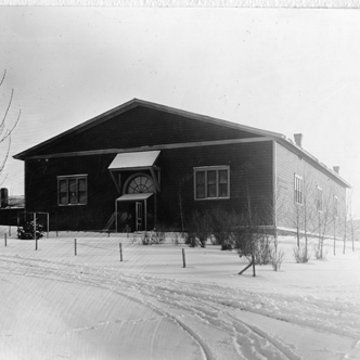 Drill Hall is a single-level rectangular building with a pitched roof. The building is surrounded by snow and young trees.