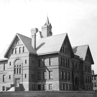 Montana Hall pictured at an angle. The four-story building is made of brick over a base of stone. Montana Hall has a unique roof line with two pitches on the front and back of the building and one pitch on both the left and right sides. Three chimneys can be seen from this angle.