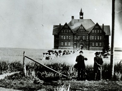 Two men wearing suits and hats stand in front of a wooden fence while women walk toward Montana Hall, a large bell-towered college administration building, through a field of tall grass on Montana State University's campus in the late 1800s.