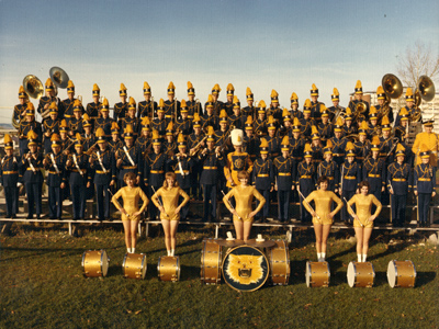 The Montana State University Marching Band stands in four rows on the bleachers on a field posing for a photo. They are wearing blue uniforms. Five majorettes stand in front of the bleachers next to drums positioned on the grass. The majorettes are wearing gold uniforms. The center drum has a painted bobcat head on it. The photo was taken between 1960 and 1970.