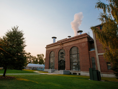 A square brick building with long arched windows on a sunny summer morning. As the whistle blows, a puff of steam is released from a vent pipe on the roof to announce the first day of fall semester classes.
