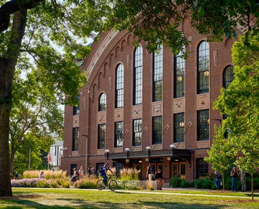 Romney Hall, a four-story rectangular brick building with a curved roof, under the shade of large trees in the summertime.