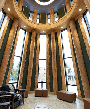 Light filters through the windows into a circular room called the drum room in MSU's American Indian Hall. The walls are made of tall wooden beams and the floor is made of concrete and stone. There are two chairs with patterned fabric and seven square leather stools pictured.