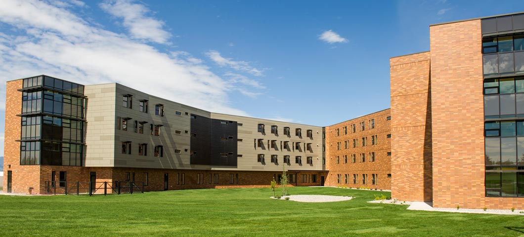 The exterior of Yellowstone Hall is made of grey metal paneling, orange brick and dark tinted windows. Yellowstone Hall is in the shape of the letter “Y”. This photo shows the intersection of the two wings. Inside the intersection is a large grassy area with newly planted trees.