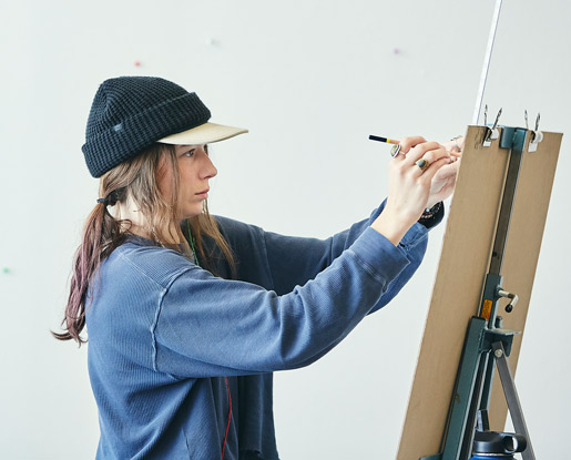 A contemporary, colored photo shows a student standing in front on an easel using a pencil and a long skinny ruler. She is wearing a baseball cap with a beanie overtop of it, has her hair in a ponytail and a navy sweatshirt. The angle of the photo does not show us what she is drawing.