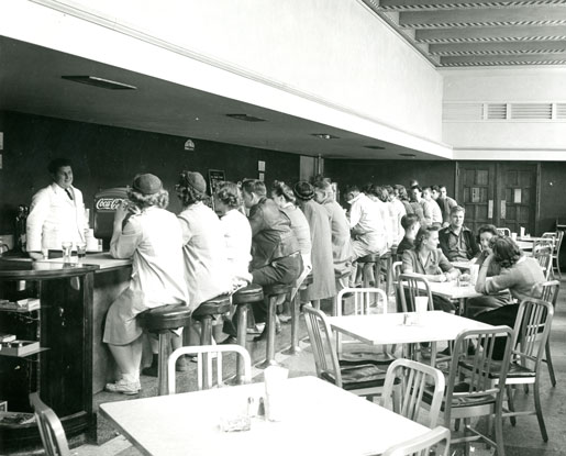 A black-and-white photo shows students enjoying a meal in a campus dining area. The room is long and narrow. Some students are shown sitting up at a long bar on barstools talking to a man behind the bar. There is a Coca-Cola machine beside the man. Other students are pictured talking among each other at tables in the center of the room.