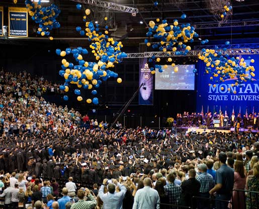 A contemporary, colored photo shows blue and gold balloons falling from the ceiling of the Brick Breeden Fieldhouse, the basketball arena on Montana State University's Bozeman campus. Graduating students stand in rows while raising their hands in celebration while parents, families and friends celebrate in the stands. A large Montana State University “M flame” logo is displayed behind the stage on a blue curtain.