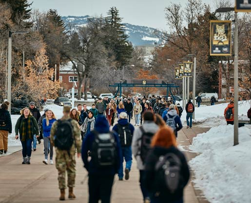 Students walk to class in the 2020s on MSU's Centennial Mall. Students walk east on the sidewalk and mountains can be seen in the background. The mountains are framed by Aasheim Gate, a decorative raised structure made of blue metal that spans the sidewalk and is topped with a polished stone slab and an artistic rendering of the letters “MSU.”