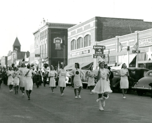 A black-and-white photo shows an MSU homecoming parade on Bozeman's Main Street in the late 1930s. Old-fashioned cars line the street, along with brick buildings bearing signs of various businesses, including the Rocking R Inn. In the foreground, female students from MSU's drill team lead the marching band down Main Street, twirling batons and wearing white knee-length skirts and sweaters.