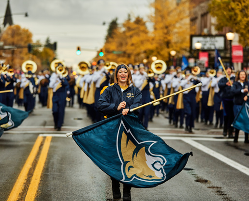 A contemporary, colored photo shows a female member of MSU's color guard spinning a flag with Montana State's Bobcat athletic logo on it. She is wearing a ear wrap with the block M logo embroidered on it, a navy hooded zip-up jacket and pants. MSU's marching band is following behind her down Bozeman's main street. Gold fall foliage towers behind them.