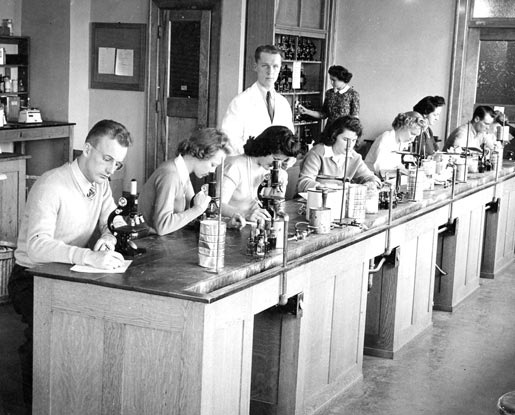 A black-and-white photo shows students in a lab working with microscopes during Montana State College's high school week. A professor stands in the background of the image wearing a white coat and tie.