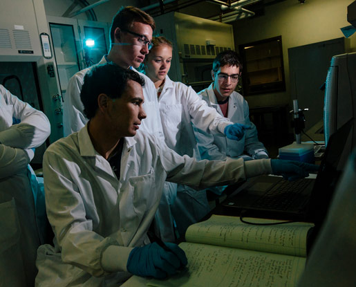 A contemporary, colored photo shows students wearing white lab coats. They are looking and pointing at a computer screen. One student in the center of the image is writing in his notebook while pointing at the computer. There is a microscope in the background of the image.