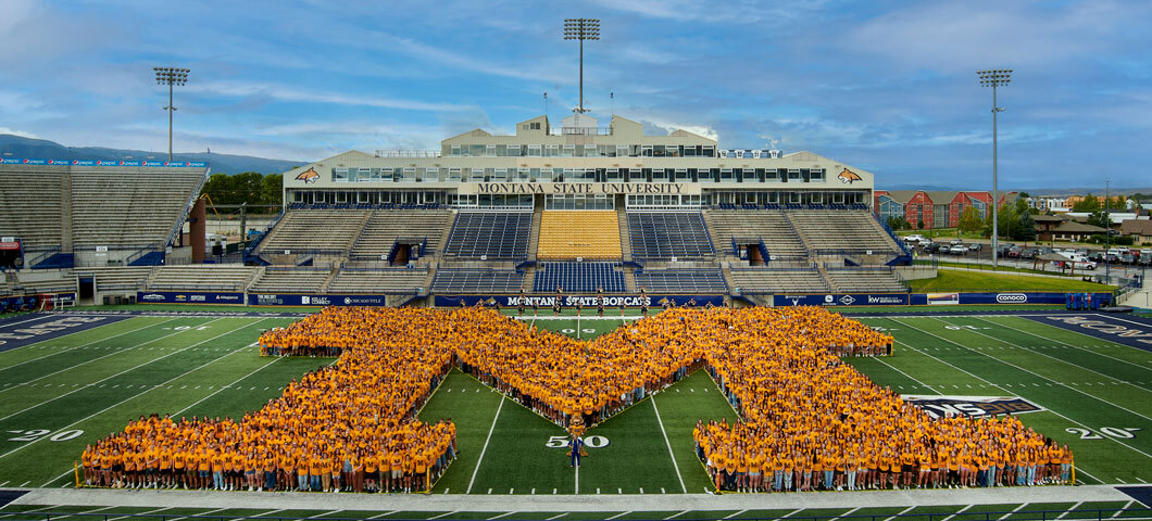 Members of Montana State University's incoming class of 2024 stand in the shape of the letter “M” on the football field in Bobcat Stadium as part of a yearly tradition. The students are all wearing matching gold shirts.