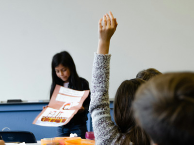 Student raising their hand, with a student doing a presentation in the background